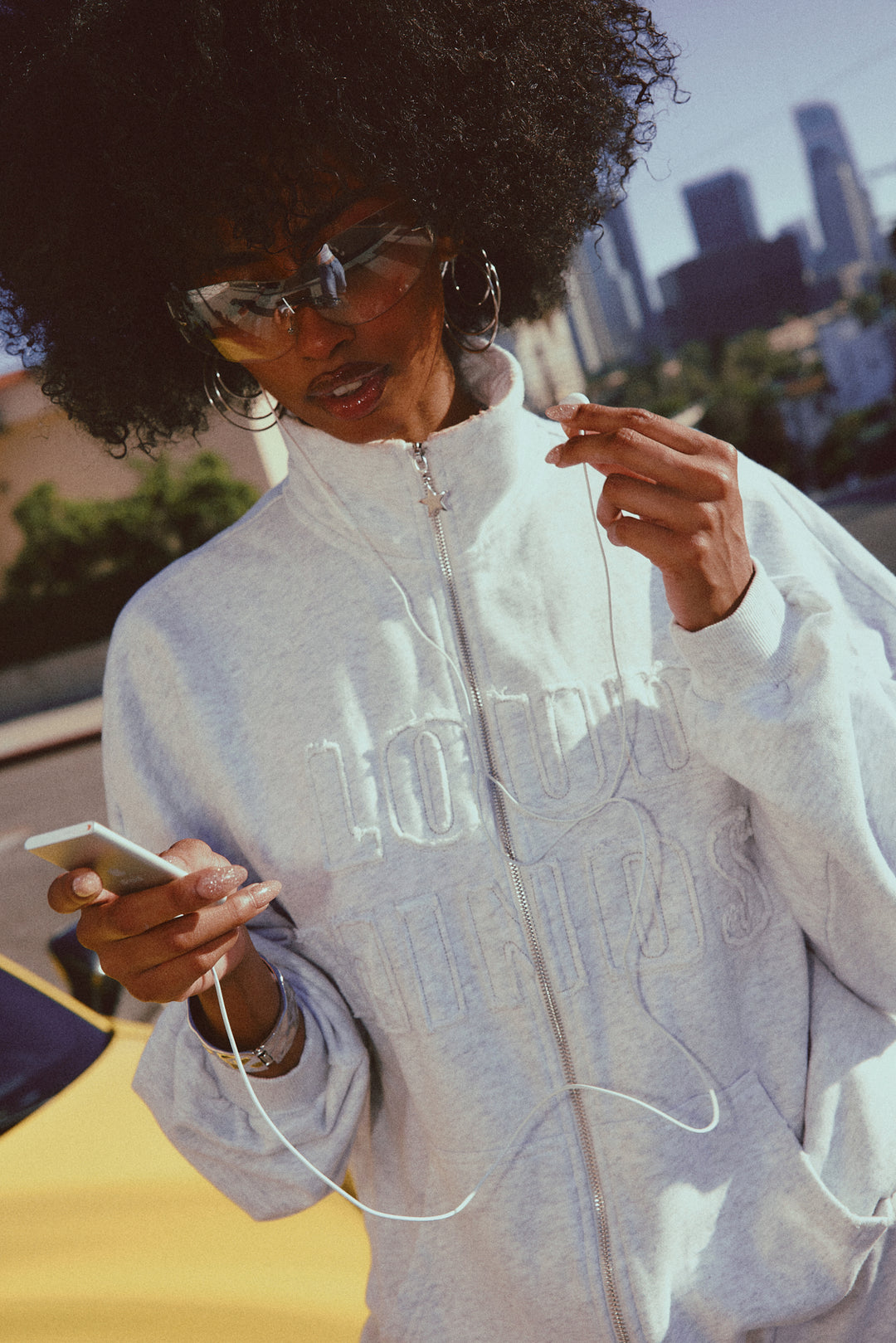 Person wearing a Heather Grey outfit jacket with text, using a phone outdoors with buildings in the background