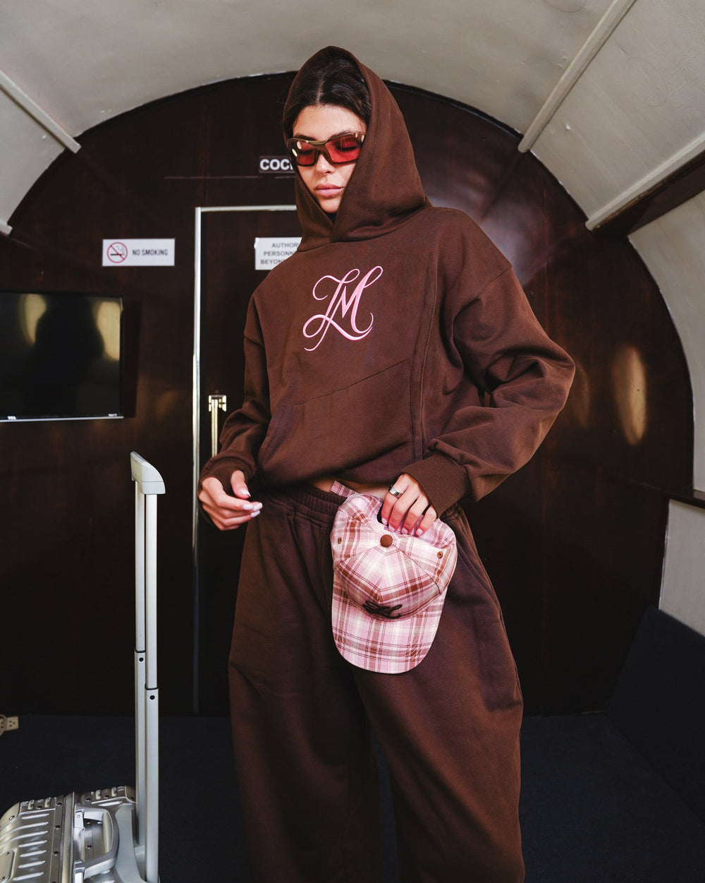 A person wearing a brown hooded sweatshirt and matching sweatpants stands in a private jet. The hoodie has a large pink monogram 'LM' embroidery on the chest, and the person is holding a pink plaid cap.
