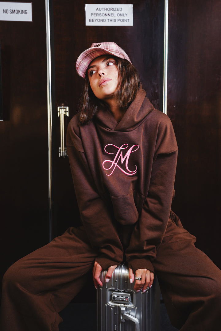 A woman wearing a brown matching hoodie and sweatpants set with a pink plaid cap sits on a silver metal case in front of dark doors. The hoodie features a large pink cursive &