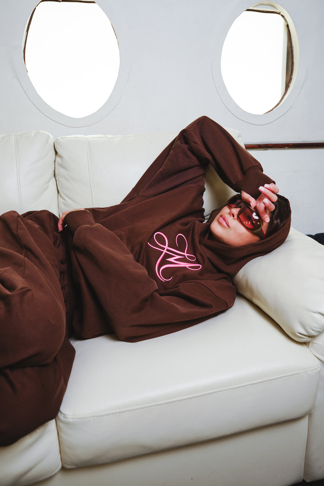 A person wearing an oversized brown hoodie and matching brown pants with a pink embroidered logo, reclining on a white sofa in front of circular window openings.