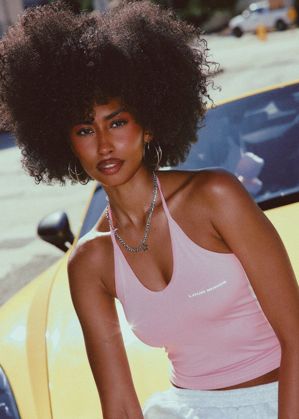 Woman in a pink halter tank top sitting next to a yellow car on a street.