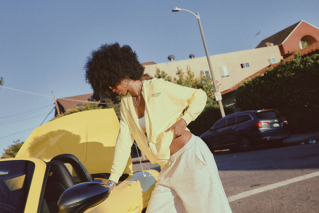 A woman with an afro hairstyle wearing a pale yellow zip-up hoodie and white sweatpants leans over a bright yellow convertible car in an outdoor street setting.