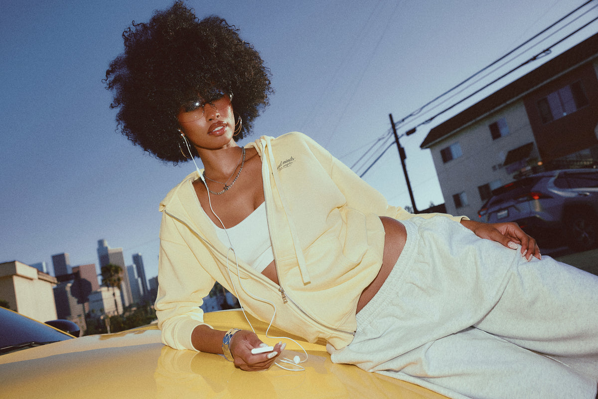 A woman with a large dark afro lies on the hood of a yellow car outdoors, wearing a pale yellow zip-up hoodie, white tank top, and white sweatpants. She has white wired earphones in and the background shows a city skyline and buildings under a blue sky.