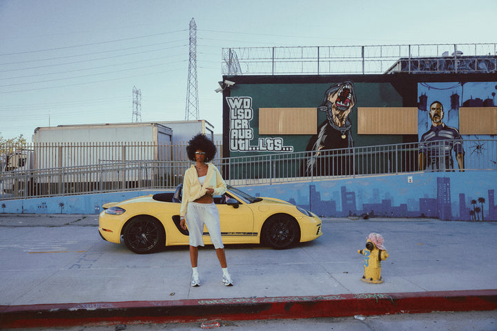 A person stands in front of a bright yellow sports car parked on a Los Angeles street, with graffiti-covered walls and urban surroundings in the background.